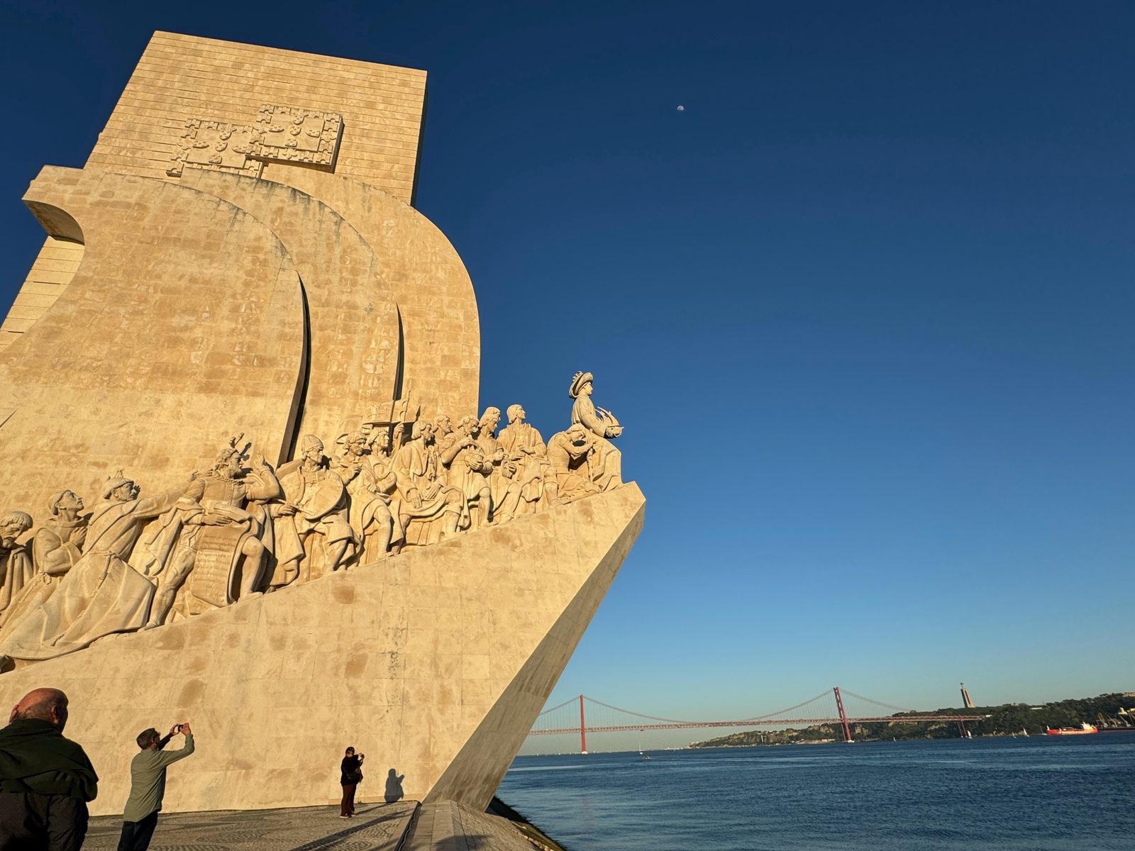 The Padrão dos Descobrimentos monument in golden light beside the Tagus River, with the 25 de Abril Bridge beyond
