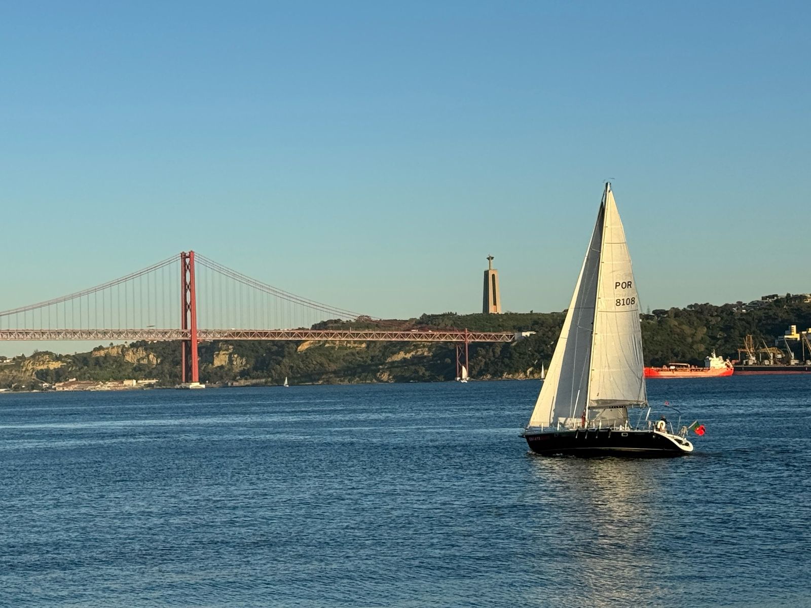 A sailboat on the Tagus River in front of the 25 de Abril Bridge and the Cristo Rei statue
